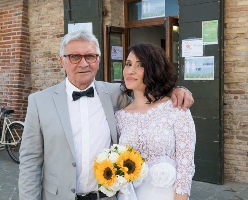 Fotografo-matrimonialista con sposa e il babbo che la tiene vicina con il braccio. Le mani della sposa tengono un bouquet di girasoli